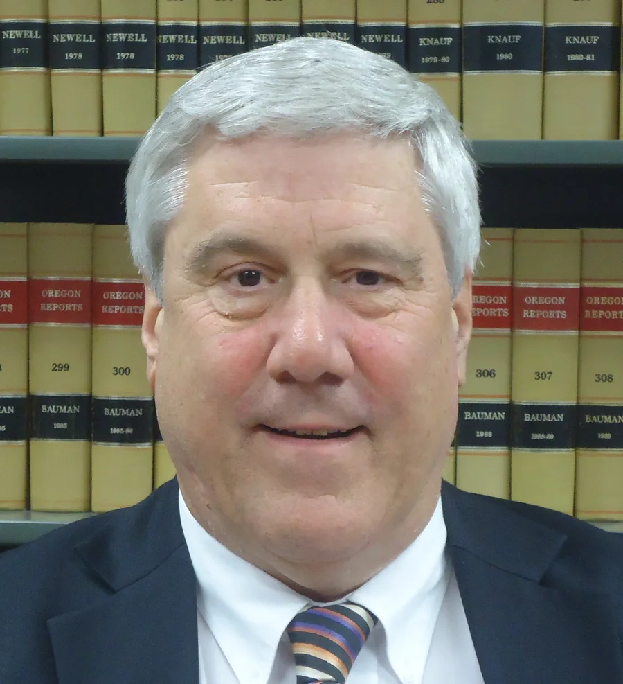 Kent Hickam, white-haired, in a navy suit and striped tie, in front of shelves of tan Oregon Reports volumes.